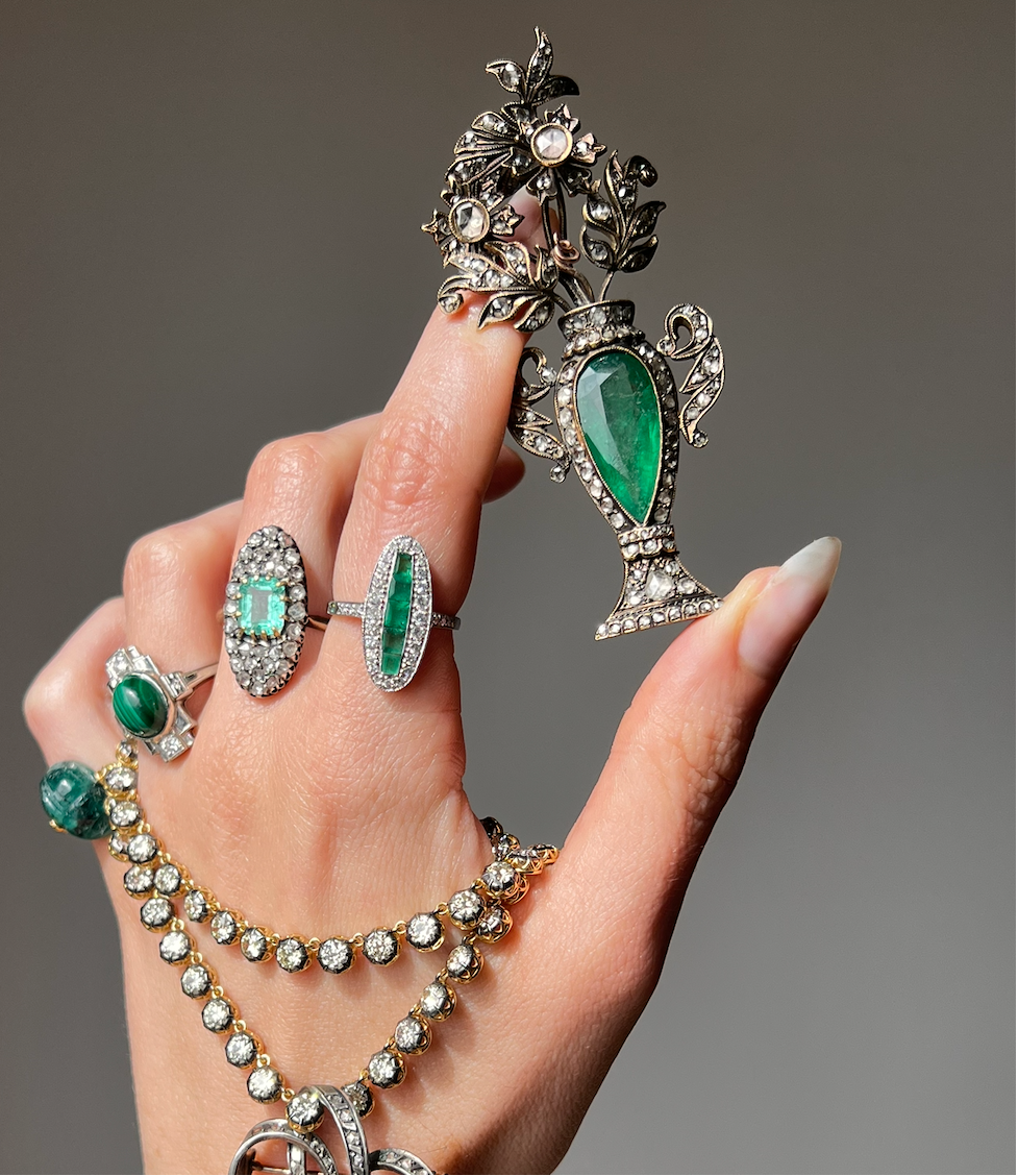 Late 18th Century Giardinetti Brooch with Large Emerald and Rose Cut Diamonds, held by a hand with an art deco emerald and diamond ring with calibre cut emeralds, a massive emerald and diamond Navette ring from the Napoleon III period, a malachite art deco ring with diamond accents, and an emerald scarab ring, draped with a diamond riviere necklace of 18", also known as a tennis necklace. 