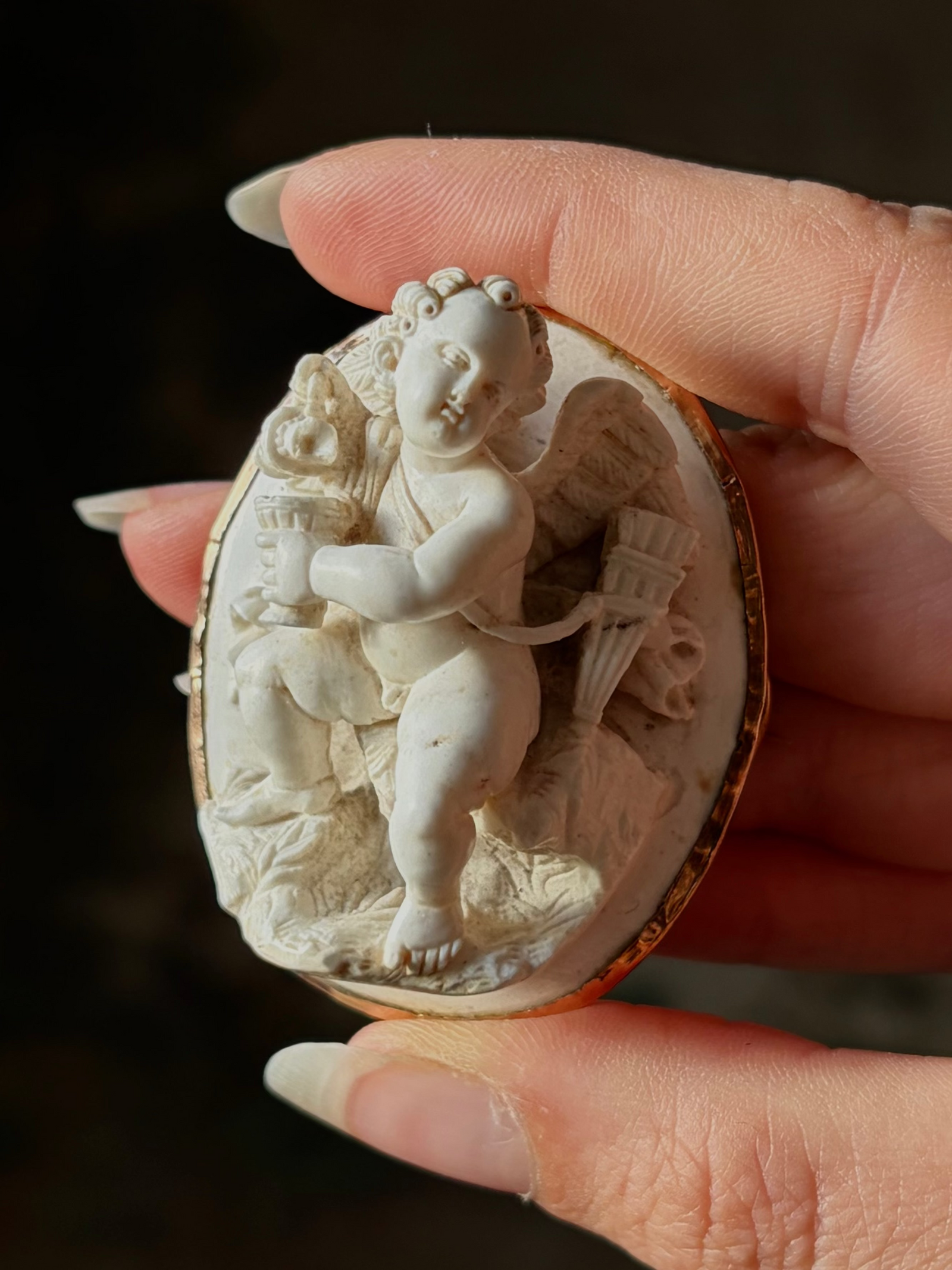 Antique white lava cameo of Cupid pouring wine, c.1860, in a 14ct gold frame, held between fingers.